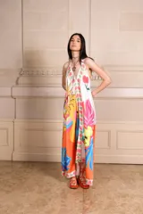 Vestido largo de seda con estampado vibrante y multicolor que incluye motivos frutales y caras sonrientes. Diseño de solera con escote halter anudado al cuello y falda amplia.