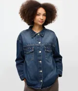 Camisa de jean estilo overshirt con corte oversized, cuello clásico y mangas largas. Presenta cierre frontal con botones, dos bolsillos delanteros con solapa y aplicación de tachuelas metálicas en el cuello y la parte superior del pecho.
