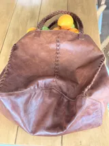 Bolso tipo tote de cuero color marrón, con diseño artesanal que presenta detalles de costuras trenzadas en los laterales y en el centro. Cuenta con dos asas superiores trenzadas y un tamaño amplio.