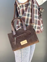 Cartera bandolera de cuero color marrón chocolate, con forro de tela y herrajes dorados. Tiene doble asa de mano y correa larga regulable y desmontable. Incluye bolsillo frontal con cierre y bolsillo interno.