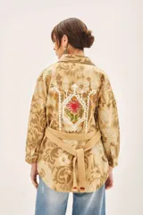 Campera de corte recto confeccionada en gabardina de algodón, con diseño estampado y detalles bordados. Presenta cuello camisero, cierre frontal con botones, bolsillos con solapa en el pecho y lazo ajustable en la cintura con terminaciones bordadas.