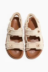 Sandalias planas de sarga de algodón en color beige con terminación desflecada. Cuentan con tiras ajustables con detalle metálico dorado y cierre autoadherente, además de una tira ajustable en el talón. La plantilla es preformada en tono marrón.