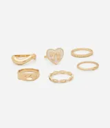 Kit de seis anillos dorados de metal reciclado. Incluye un anillo con forma de corazón esmaltado en blanco con un diseño de moño, un anillo con forma de onda, y cuatro anillos finos con detalles texturizados.