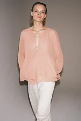 Blusa de viscosa color rosado con cuello redondo, manga larga abullonada con puños elásticos y cierre frontal con lazadas. Presenta un diseño con volantes en el bajo y corte asimétrico.
