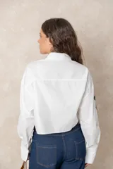 Camisa blanca de algodón, de corte corto y silueta holgada. Presenta cuello clásico, cierre frontal con botones y mangas largas con puños anchos. Está decorada con diversos parches y pines metálicos de estilo militar y náutico distribuidos en el frente y la manga derecha.