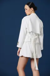 Saco corto de color blanco con solapas clásicas, mangas largas y detalle de parche rectangular con texto en el puño izquierdo. Presenta una silueta estructurada y elegante.