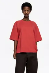 Camiseta negra de corte recto y largo medio, confeccionada en algodón orgánico. Presenta escote redondo, mangas cortas amplias con costuras raglán y detalles de cortes diagonales en el cuerpo.
