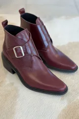 Botin de cuero color bordó, con diseño de puntera cuadrada y detalle de hebilla metálica lateral. Presenta una textura combinada en el lateral y suela de goma negra.