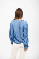 Camisa de tela liviana color azul medio, con cuello en V, mangas largas abullonadas y puños con elástico y volados.