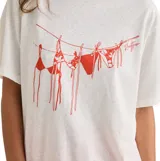 Remera de corte oversized color blanco, con cuello redondo y mangas cortas. Presenta un estampado gráfico en color rojo en el frente que ilustra bikinis colgados en una soga.