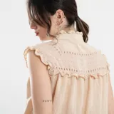 Camisa sin mangas color beige, con cuello alto con volados, detalles de encaje calado en los hombros y parte superior del pecho, y cierre frontal con botones.