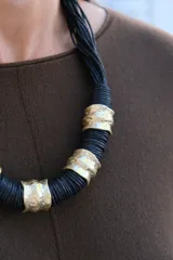 Collar de diseño artesanal compuesto por múltiples hilos de cordón negro entrelazados, intercalados con piezas cilíndricas de metal dorado con textura irregular y acabado rústico.
