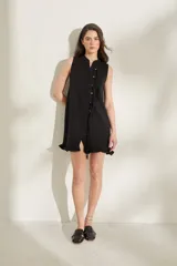 Vestido corto de punto sin mangas, color negro, con cuello redondo y cierre frontal decorado con botones y tiras colgantes. Presenta terminaciones con volados en el ruedo.