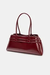Bolso de hombro tipo city en color bordó con acabado brillante. Presenta un diseño estructurado con detalle de tiras decorativas con hebillas metálicas en los laterales y doble asa de hombro.