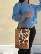 Bolso tipo matera confeccionado en cuero, con diseño combinado que presenta laterales con textura de cocodrilo y panel central con estampado animal print de vaca. Cuenta con asas largas para llevar al hombro y base reforzada.