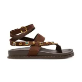 Sandalias planas de gamuza marrón, con tiras cruzadas y correa envolvente al tobillo con hebilla dorada. Incorporan tachas metálicas doradas y aplicaciones de piedras decorativas en las tiras.