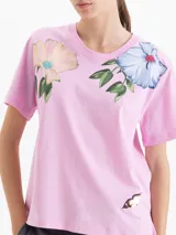 Remera de algodón con cuello redondo y mangas cortas. Presenta un diseño estampado con flores grandes en la parte superior delantera y un pequeño detalle bordado en la parte inferior.