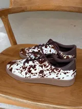 Championes urbanos con diseño de estampado animal print tipo vaca, confeccionados en cuero con textura de pelo. Presentan cordones marrones, suela de goma en tono tostado y detalles en color marrón oscuro en el cuello y lengüeta.