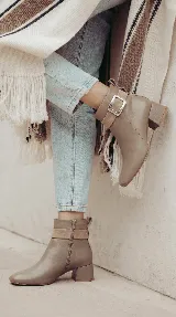 Bota corta de cuero color taupe, con diseño minimalista y taco cuadrado de 4 cm. Presenta una tira decorativa en el tobillo con una hebilla metálica rectangular y detalle de gamuza en la zona de la hebilla. Cuenta con cierre lateral interno.
