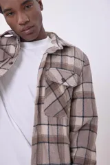 Sobrecamisa de corte holgado con diseño a cuadros en tonos beige y marrón, confeccionada en tejido de textura suave. Presenta cuello clásico, cierre frontal con botones, dos bolsillos con solapa en el pecho y puños abotonados.