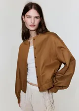Chaqueta de diseño corto en tono tostado, confeccionada con mezcla de lyocell, algodón y lino. Presenta cuello redondo, mangas largas abullonadas, elásticos en los laterales y cierre frontal de botonadura visible.