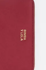 Billetera mediana de estructura rígida en color marrón topo, con cierre perimetral de cremallera y tirador decorativo de cordón. Presenta el logo de la marca aplicado en letras metálicas doradas en el frente.