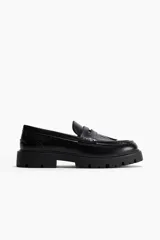 Mocasines de cuero sintético negro con suela chunky, detalles de flecos en el empeine, tira con aplique metálico dorado y costuras pespunteadas.