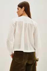 Camisa de algodón blanco con textura de pequeños puntos en relieve, cuello redondo con cierre de botones oculto, detalles de alforzas verticales y encaje en el frente, y mangas largas con puños elásticos.