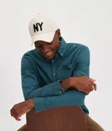 Gorra de béisbol de seis paneles, color beige claro, con visera curva y bordado frontal en negro con las letras "NY". Cuenta con cierre strapback.