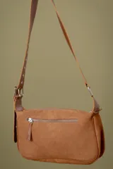 Cartera tipo bandolera de cuero color marrón, con diseño rectangular y cierre superior. Presenta detalles de tachas metálicas circulares en los bordes laterales e inferiores, y una correa larga ajustable con hebilla metálica. Incluye una placa metálica con el logo de la marca en la parte frontal.