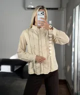 Camisa blanca de estilo romántico con cuello alto, cierre frontal de botones y detalles de encaje y alforzas verticales. Presenta mangas largas con puños decorados con encaje.