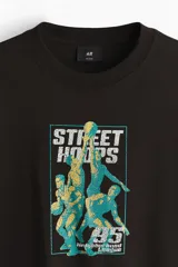 Remera de algodón marrón oscuro con cuello redondo y mangas cortas. Presenta un estampado frontal con gráficos de básquetbol y texto 'Street Hoops 95'.