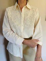 Camisa de manga larga en color beige, con cuello clásico y cierre frontal de botones. Presenta un delicado aplique de encaje tipo crunie en la parte superior delantera, aportando textura y un detalle artesanal al diseño.
