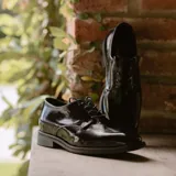 Zapato mocasín de cuero vacuno color negro, con cordones y punta redonda. Tiene taco de madera foliado en cuero de 2,5 cm y entre suela de goma de 1 cm.