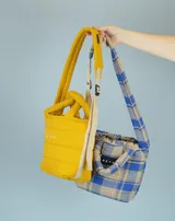 Bolsito tipo tote bag de diseño acolchado con doble asa corta y correa larga ajustable. Disponible en dos versiones: una lisa en color amarillo con logo estampado y otra a cuadros en tonos azul y beige con etiqueta de marca.