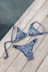 Bikini azul con efecto brillante, compuesto por un top de triángulos fijos unidos por una argolla, con breteles regulables y lazo en la espalda. El bottom es vedetina con argollas en la cadera.