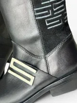 Bota de caña alta en cuero negro con punta redonda y taco bajo. Presenta un panel lateral de gamuza con bordado geométrico en hilo gris, herraje metálico decorativo en el empeine y cierre interno. El diseño cuenta con una boca asimétrica y forro interior de cuero.