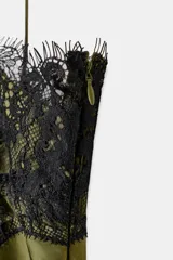 Vestido midi de satén color verde oliva, con escote redondo y tirantes finos. Presenta aplicaciones de encaje negro en el escote y en el bajo, con un diseño asimétrico.