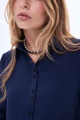 Camisa blanca de manga larga con cuello clásico y cierre frontal de botones negros en contraste. Presenta un bolsillo frontal en el pecho y un diseño de líneas simples y corte recto.
