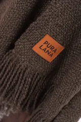 Ruana de tejido de punto color marrón chocolate, con diseño envolvente, cuello en V y detalle de flecos en el ruedo. Incluye etiqueta de cuero sintético con el logo de la marca en la parte frontal.