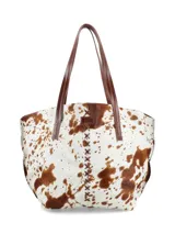 Bolso tipo tote de piel de vaca, con estampado moteado en blanco y marrón. Presenta asas de cuero marrón oscuro y costuras decorativas en forma de X en el centro frontal.