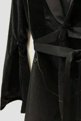Blazer tipo smoking de terciopelo negro con acabado brillante y detalles estampados en dorado. Presenta solapas clásicas, cierre frontal cruzado mediante un lazo y mangas largas con cierres incorporados. El diseño es entallado y sofisticado.