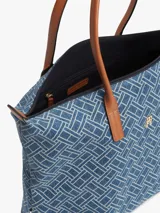 Cartera tote Tommy Hilfiger confeccionada en tejido denim azul con estampado geométrico de monogramas. Cuenta con asas superiores de cuero sintético marrón, cierre superior de cremallera y herrajes metálicos dorados con el monograma de la marca en el frente.