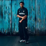 Camiseta de fútbol de manga corta en color negro, con cuello en V. Presenta el logo de la marca Corteiz en el pecho, junto a un escudo con los colores de la bandera italiana y una estrella dorada. El nombre de la marca aparece estampado en grande en el centro del frente.