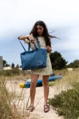 Bolso tipo tote confeccionado en tela denim de color azul claro. Presenta un diseño amplio con costuras visibles, doble asa de hombro y un detalle bordado circular en color rojo en la parte inferior.