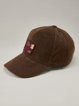 Gorra tipo baseball de pana marrón con parche rectangular bordado en color bordó y detalle de logo dorado.