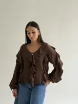 Blusa blanca de corte holgado con escote en V, decorada con volados frontales y en las mangas. Presenta mangas largas con puños elásticos y cierre frontal con botones.