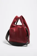 Bolso de mano mediano de piel vacuna, color negro, con estructura blanda y forma de cubo o 'paper bag'. Posee asas de mano cortas y una correa ajustable y extraíble de tela azul marino. El logo de Bimba y Lola está grabado en la parte inferior frontal.