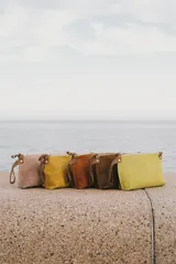 Sobre de mano confeccionado en cuero agamuzado con cierre metálico superior y correa lateral pequeña. Presenta un diseño minimalista con detalles de remaches metálicos en los extremos del cierre.