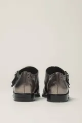 Zapato tipo monk strap con aberturas laterales, confeccionado en cuero vacuno metalizado color bronce. Presenta detalles de perforaciones estilo brogue en la puntera y el talón, y doble hebilla metálica en el empeine.